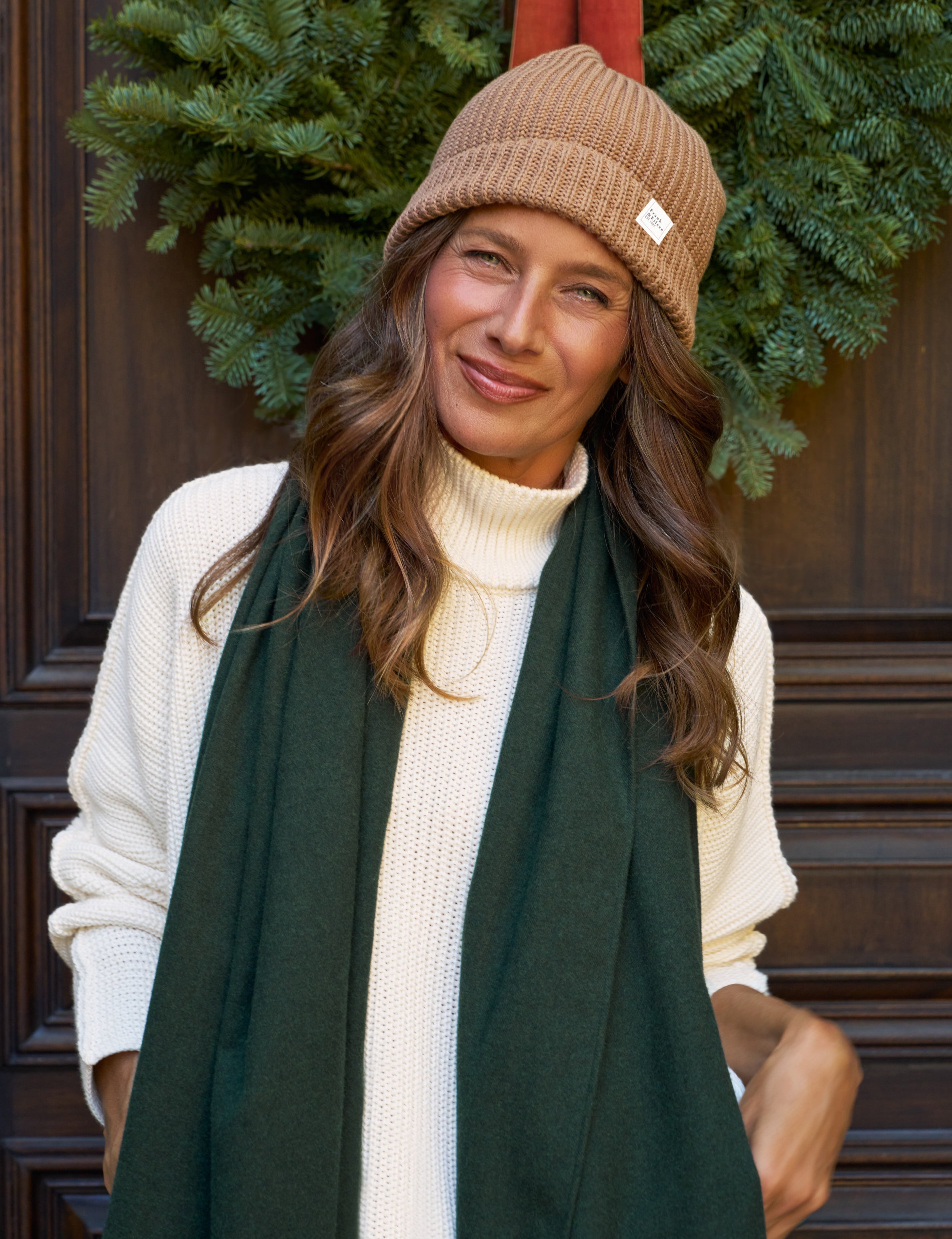 Woman wearing a brown beanie and green scarf in front of a wooden door with greenery.