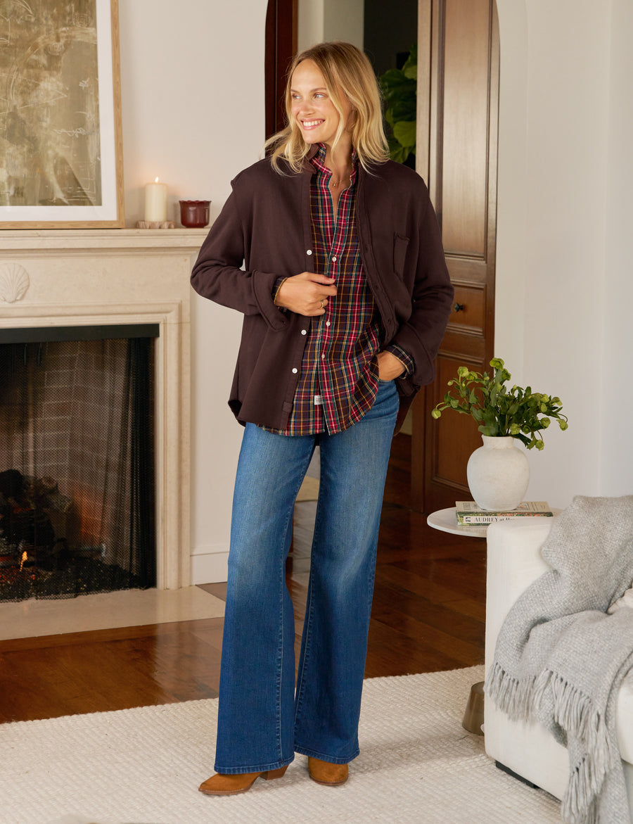 Woman standing in a living room wearing a brown jacket, plaid shirt, and blue jeans.