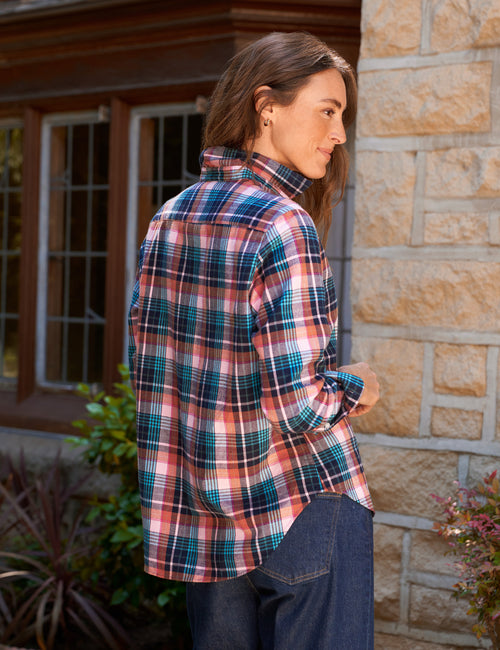 A woman wearing a long-sleeve plaid button-up shirt in pink, blue, and white, paired with dark blue jeans, standing outdoors next to a stone wall., alternate view