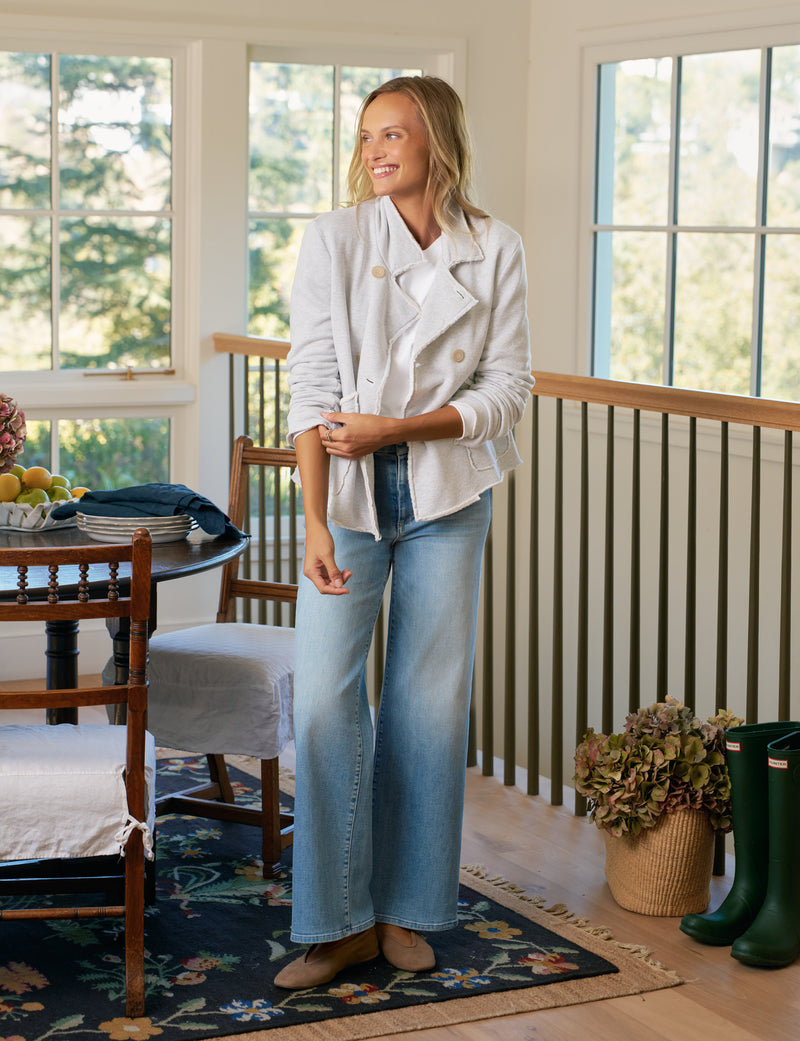 Woman standing in a room with large windows, wearing a light jacket and jeans.