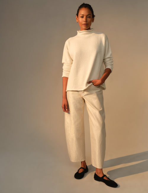 A model stands against a warm beige studio background wearing a cream-colored knit mock-neck sweater and matching barrel-leg cropped trousers, paired with black flat shoes., view 5