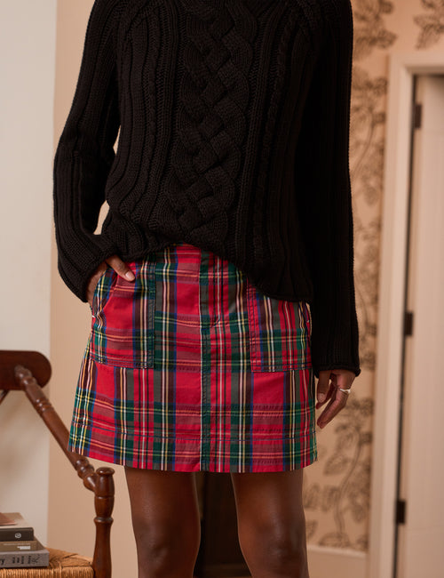 A person wearing a black cable-knit sweater and a red tartan plaid mini skirt with green and yellow stripes, standing indoors., front view