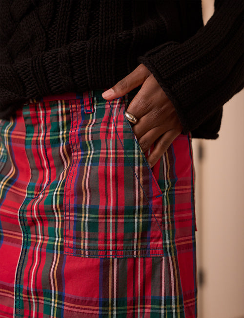 A close-up of a person wearing a red tartan plaid skirt with green, yellow, and white stripes, paired with a black ribbed sweater. The person's hand is in the pocket of the skirt., view 3