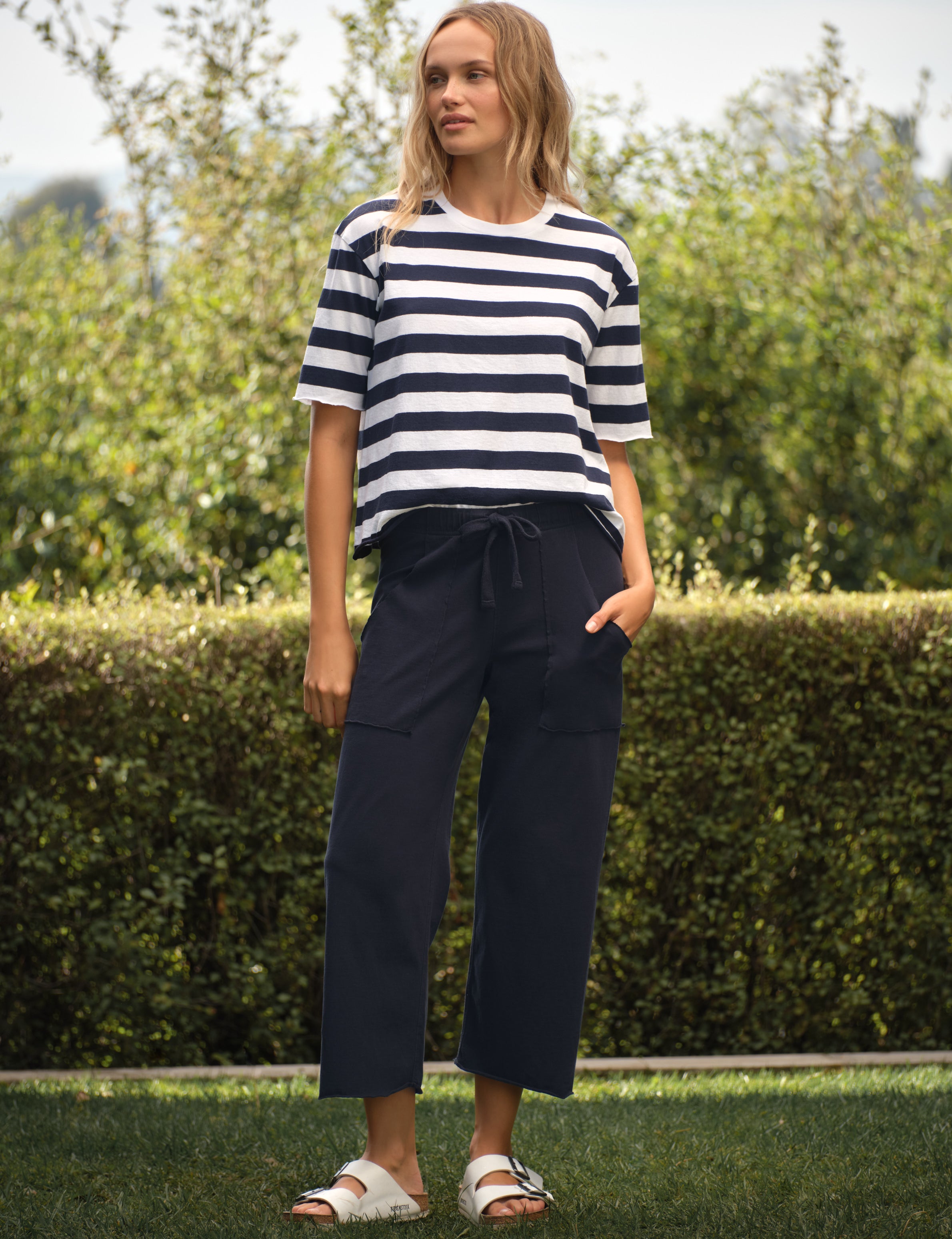 Woman wearing a striped shirt and navy pants standing outdoors with greenery in the background, view 4