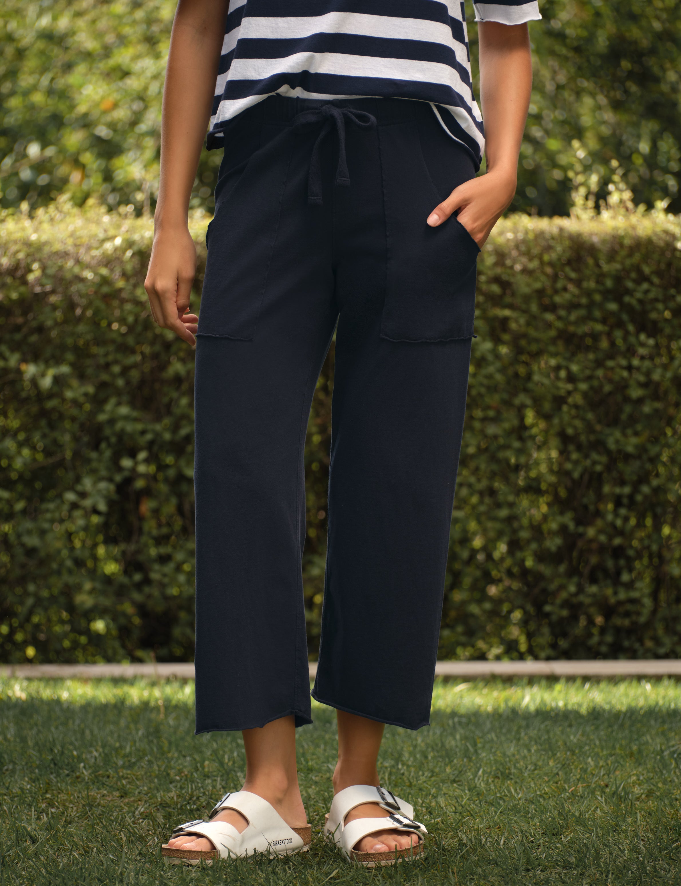 Person wearing navy pants and a striped shirt standing on grass with a hedge in the background, front view