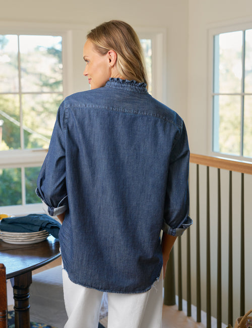 Person wearing a blue denim shirt standing in a room with large windows., alternate view