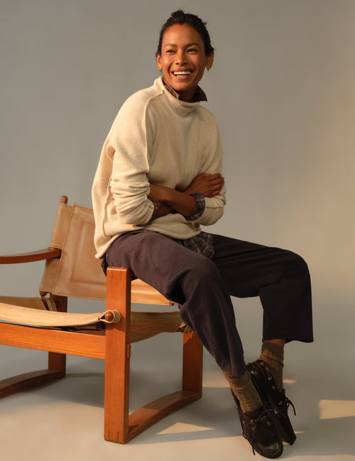 A smiling woman sits on a wooden chair with beige fabric seats, wearing a cream turtleneck sweater, dark cropped pants, brown socks, and black lace-up shoes against a plain light gray studio background., view 4