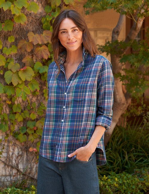 A woman wearing a blue and green plaid button-down shirt with dark jeans, standing outdoors in front of a vine-covered tree., front view