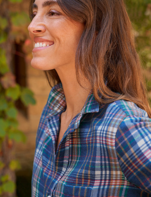 A woman in profile wearing a blue, green, and white plaid button-up shirt, photographed outdoors with soft-focus foliage in the background., view 3