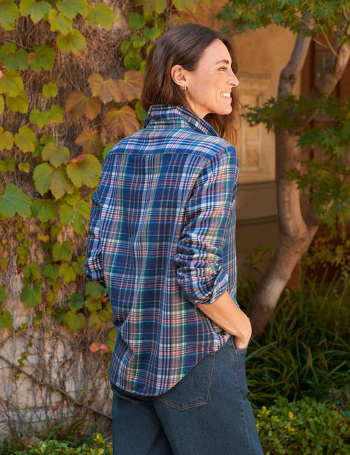 A woman wearing a blue and green plaid button-down shirt with dark jeans, standing outdoors in front of a vine-covered wall., alternate view