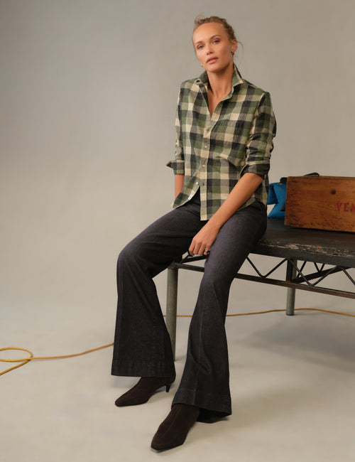 A woman sits on a bench in a studio setting, wearing a green and beige plaid button-down shirt, dark charcoal flared pants, and dark brown pointed-toe boots. A wooden crate and a blue bag are visible on the bench next to her., view 4