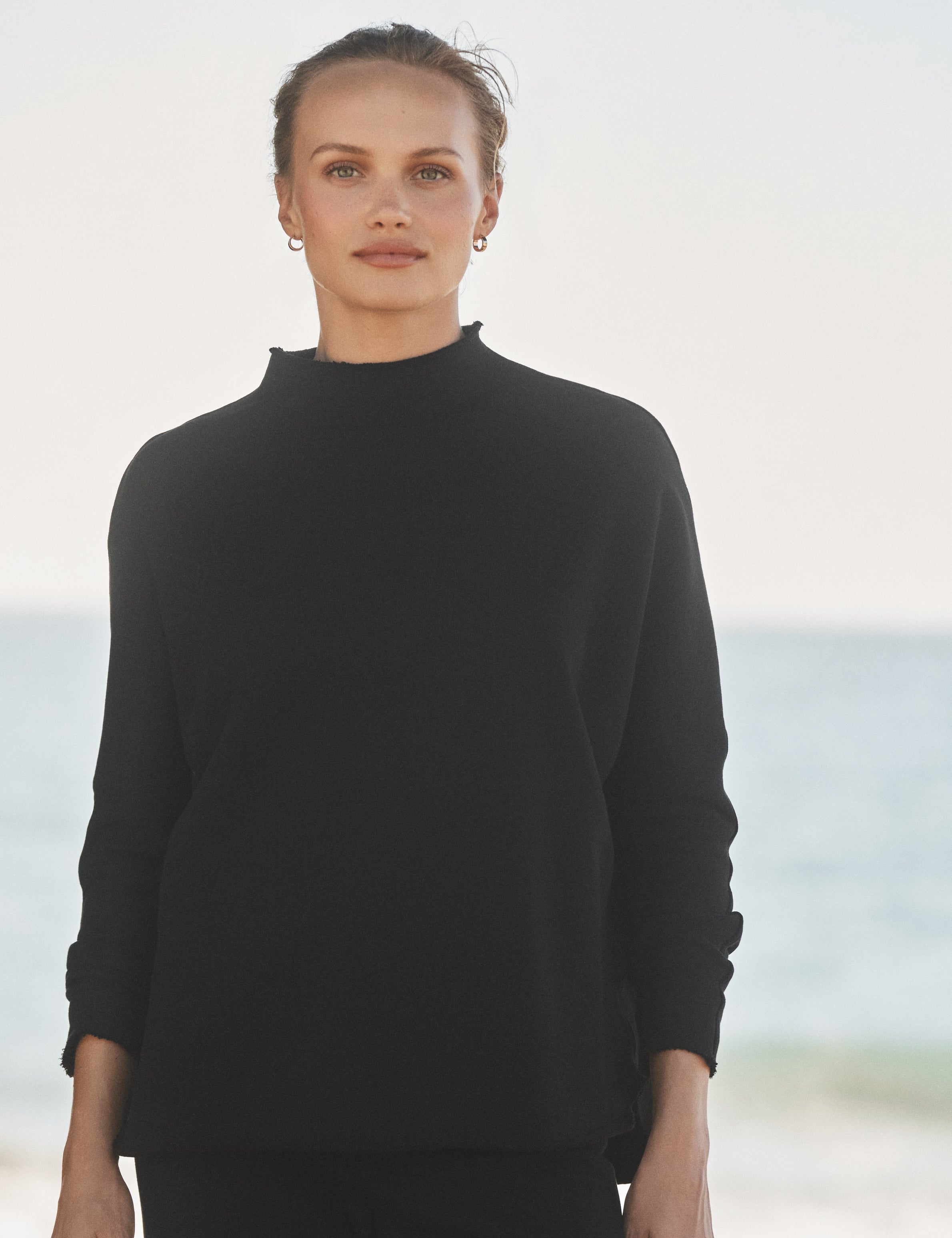Woman wearing a black sweater standing on a beach with ocean in the background, front view
