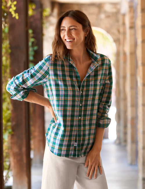 A woman smiling in a stone corridor wearing a green, blue, and beige plaid button-down shirt and light beige pants., front view