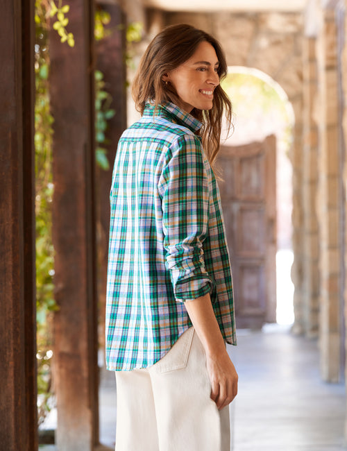 A woman wearing a green and blue plaid button-down shirt with white pants, standing in a rustic stone corridor., alternate view