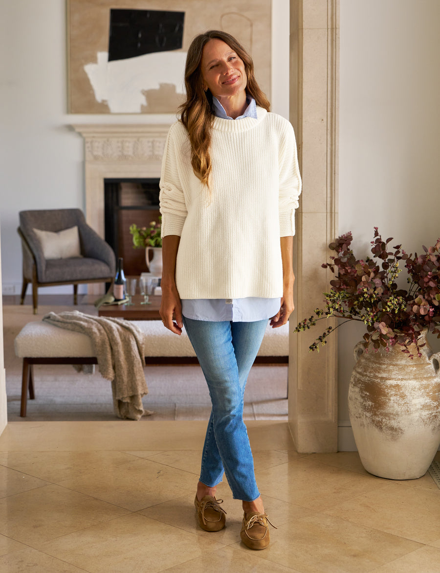 Woman standing in a living room wearing a white sweater and blue jeans.