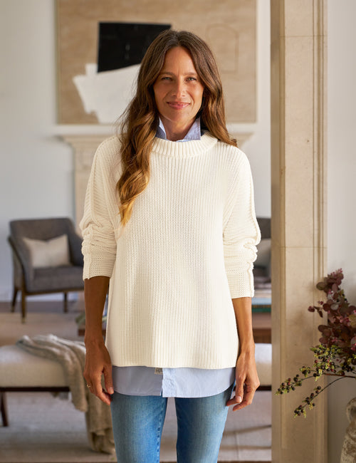 Woman wearing a white sweater over a blue shirt in a living room setting, front view