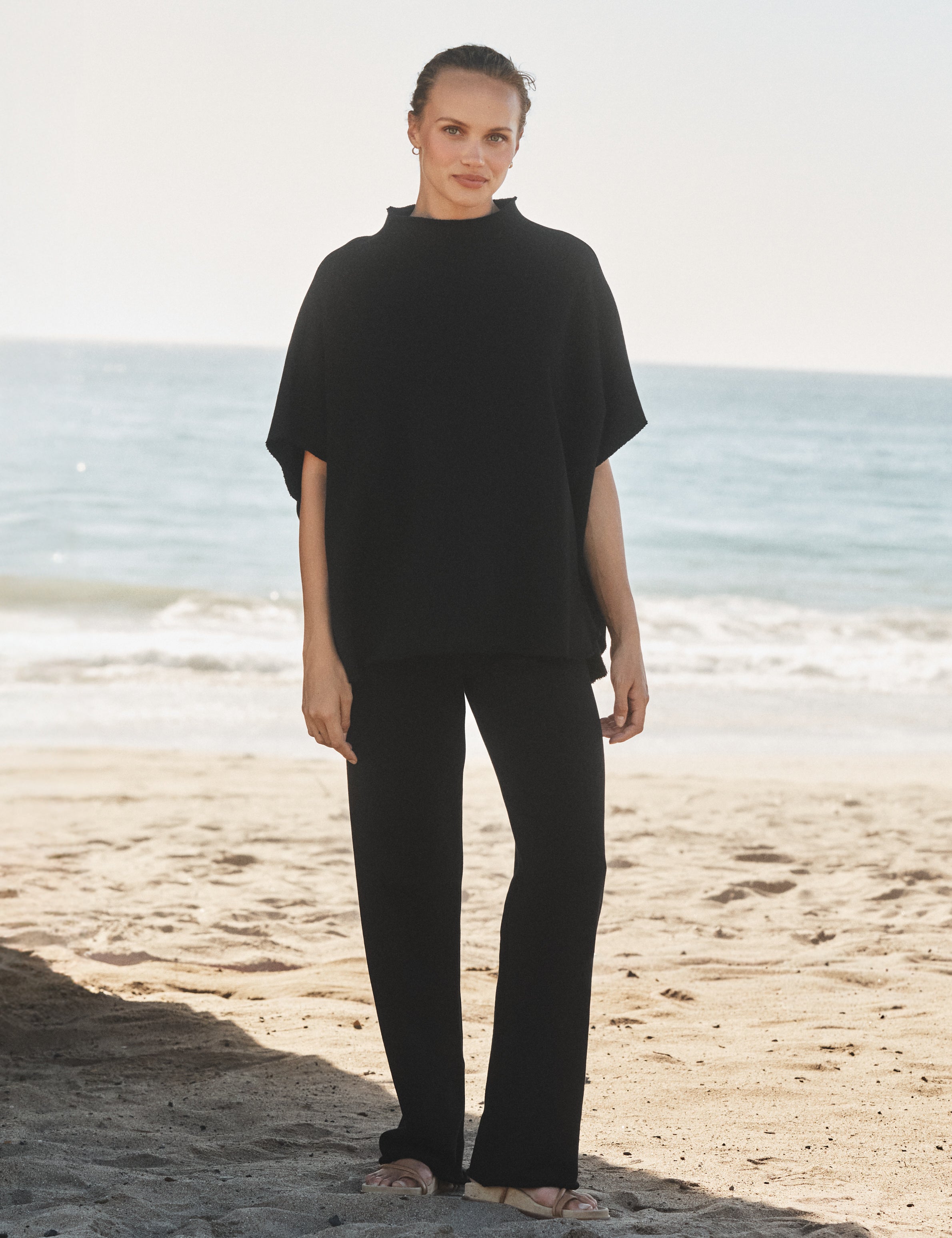 Person wearing a black capelet and pants standing on a beach with ocean in the background, front view