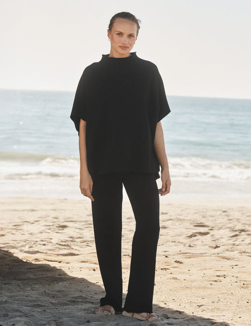 Person wearing a black capelet and pants standing on a beach with ocean in the background, front view