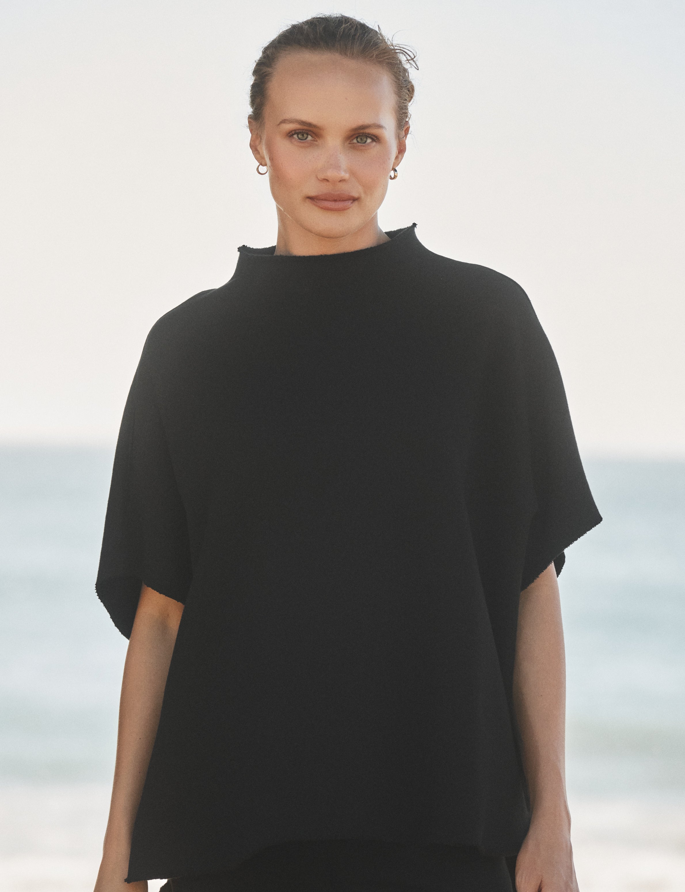 Woman wearing a black capelet with a blurred beach background, view 3