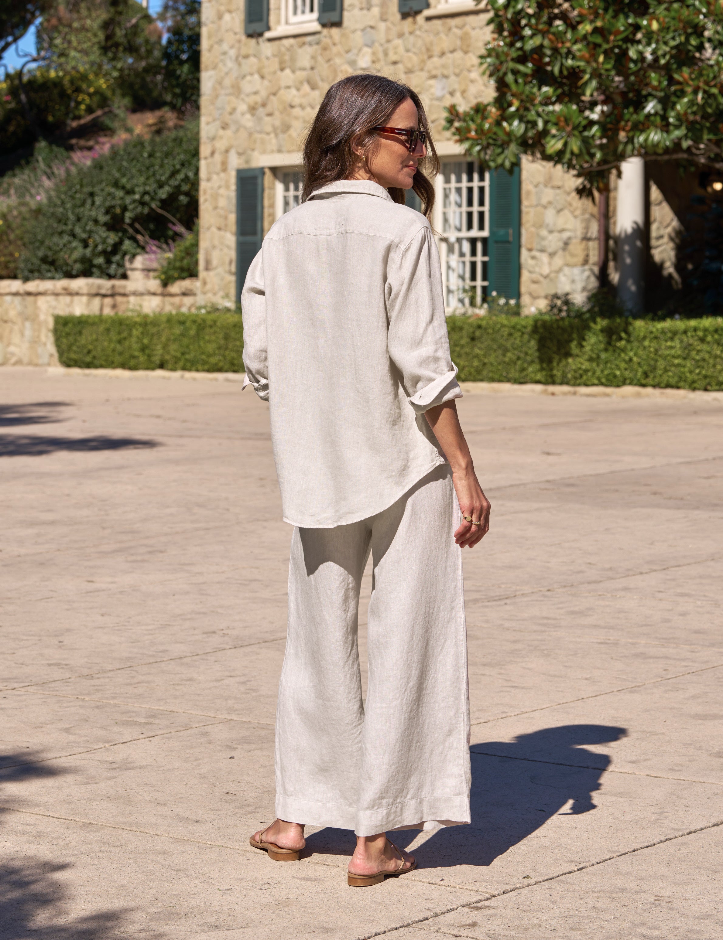 Woman in a light-colored outfit standing on a stone path with a building and trees in the background