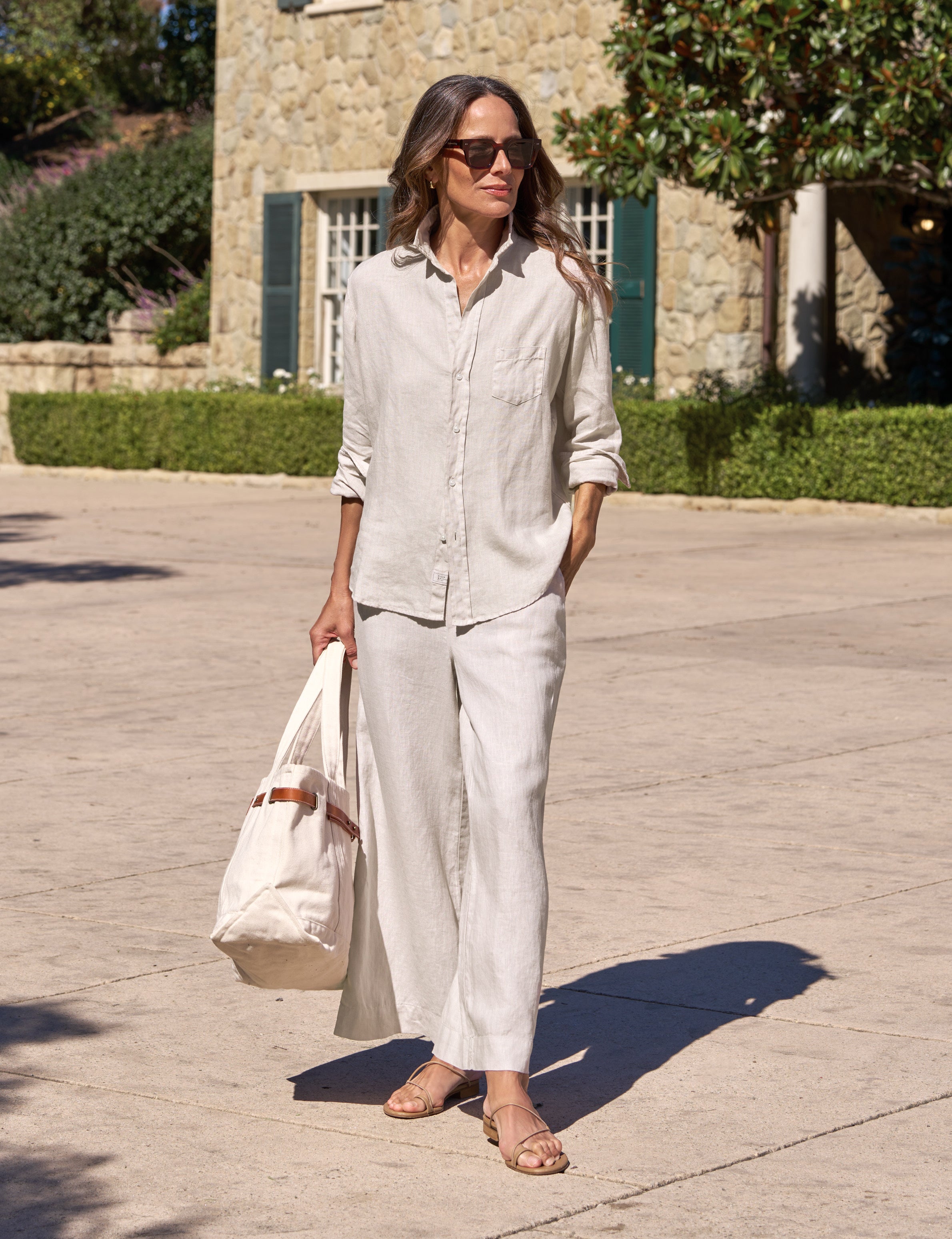 Woman in a light outfit standing outdoors with a building and greenery in the background