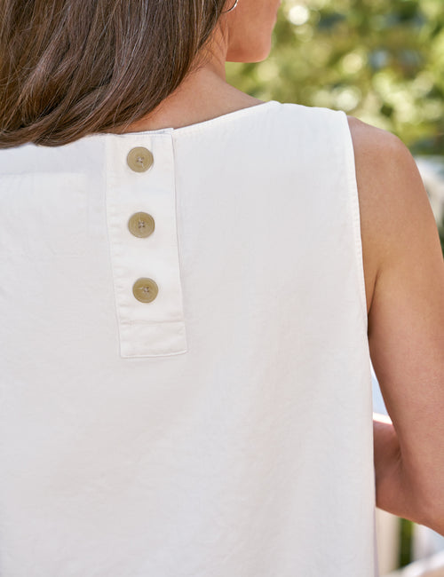 Close-up back view of a woman wearing a sleeveless white top with three gold buttons down the center back, set against a blurred outdoor background., view 3