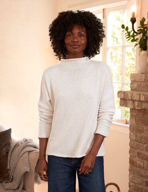 A woman with an afro hairstyle wearing a cream ribbed knit mock neck sweater and dark blue jeans, standing in a warmly lit living room next to a brick fireplace., front view