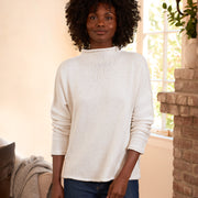 A woman with an afro hairstyle wearing a cream ribbed knit mock neck sweater and dark blue jeans, standing in a warmly lit living room next to a brick fireplace.