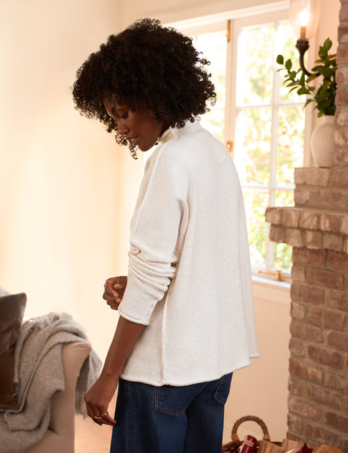 A woman with curly black hair is shown from the back wearing a cream-colored, textured knit cardigan and blue jeans in a warm, sunlit room with a brick fireplace., alternate view