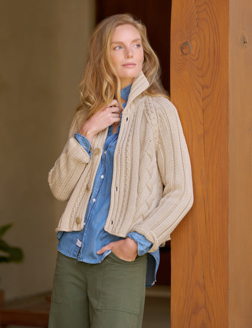 A woman wearing a cream cable-knit button-front cardigan over a blue denim shirt and olive green pants, standing next to a wooden door frame., front view