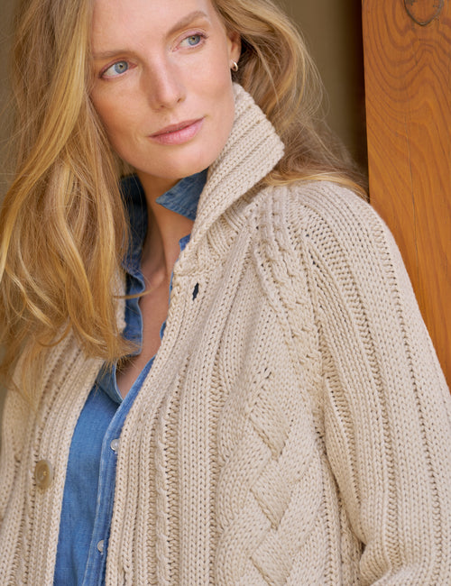 A woman wearing a cream cable-knit cardigan over a blue denim shirt, photographed in a warm indoor setting with a wooden door visible in the background., view 3