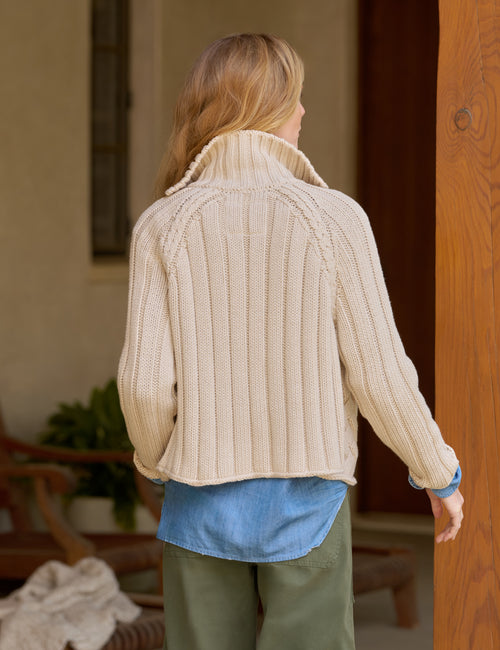 A woman shown from behind wearing a cream ribbed knit mock neck cardigan, a blue shirt, and olive green pants in an indoor setting., alternate view