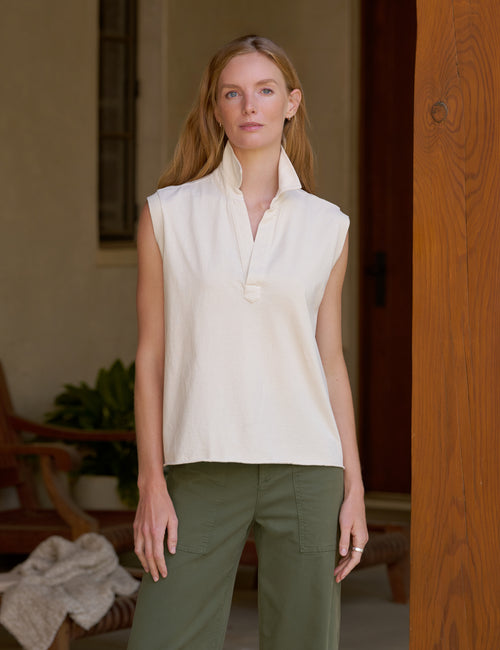 A woman modeling a sleeveless cream-colored collared top, paired with olive green pants, standing in a warm-toned indoor setting., front view
