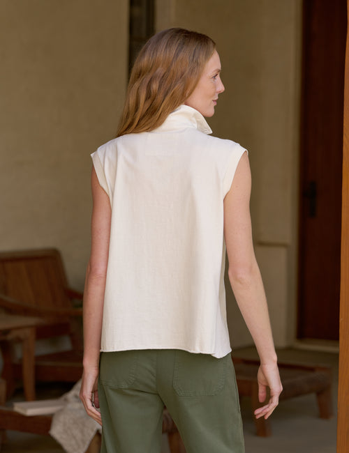 A woman shown from the back wearing a sleeveless cream-colored top and olive green pants, standing in a softly lit indoor setting with warm neutral walls and wooden furniture in the background., alternate view