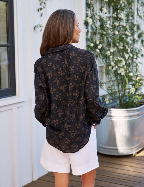 Woman wearing a black floral blouse and white shorts standing on a wooden deck., alternate view