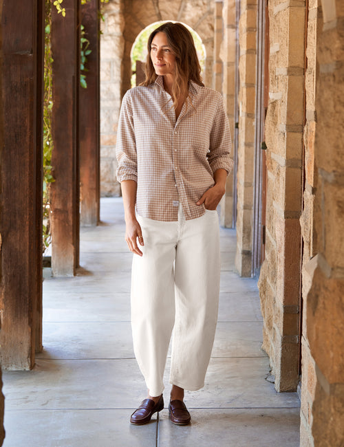 A woman stands in a stone and wood covered walkway wearing a beige and white check button-down shirt, white wide-leg cropped pants, and dark brown leather loafers., view 4