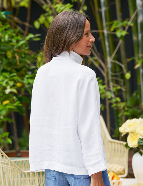 A person wearing a white washed linen shirt with a mandarin collar and short sleeves, paired with blue denim shorts, standing in an outdoor setting with greenery and a white chair in the background., alternate view