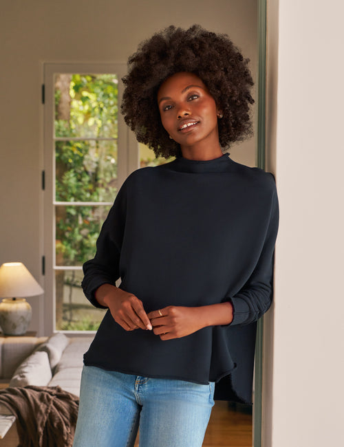 Woman wearing a navy top and blue jeans standing in a room with a window in the background., front view