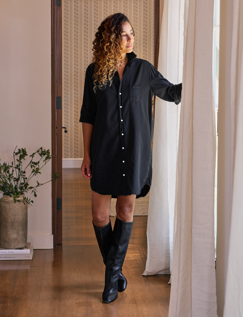Woman wearing a black dress and knee-high boots standing in a room with curtains.