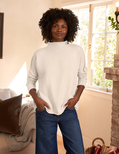 A woman with curly black hair wearing a cream-colored mock neck knit sweater and blue jeans, standing in a brightly lit living room with a window and brick fireplace., front view