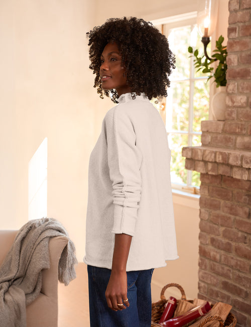 A woman with curly black hair stands in profile wearing a cream long-sleeve top and dark blue jeans in a warm, sunlit room with a brick fireplace and a window., alternate view