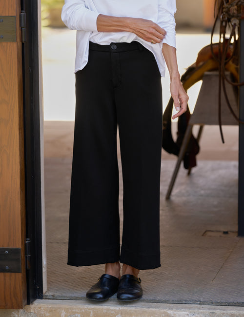 A person wearing black wide-leg cropped pants with a white button-up shirt and black loafers, standing in a doorway., view 4