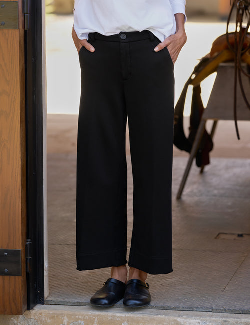 A person wearing black wide-leg cropped trousers with a white top and black loafers, standing in a doorway., front view