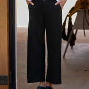 A person wearing black wide-leg cropped trousers with a white top and black loafers, standing in a doorway.