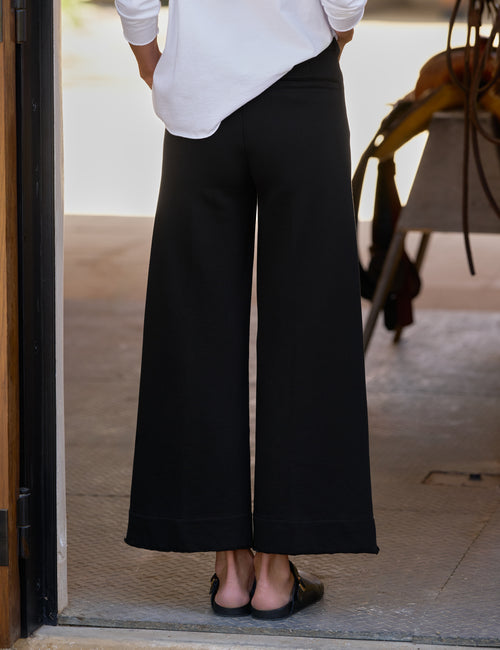 A person shown from the back wearing black wide-leg cropped pants with a white long-sleeve top and black sandals, standing in a doorway., alternate view