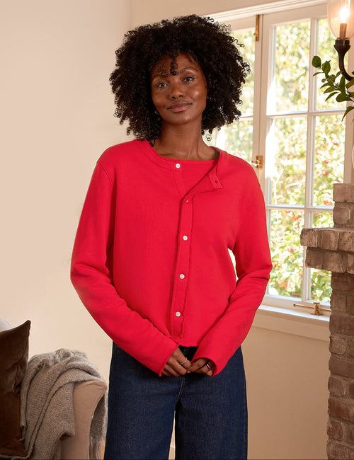 A woman stands in a cozy living room wearing a bright red button-up cardigan, dark blue wide-leg pants, and black flats. The room features a brick fireplace, a beige sofa, and a wicker basket., front view