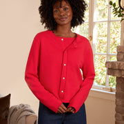 A woman stands in a cozy living room wearing a bright red button-up cardigan, dark blue wide-leg pants, and black flats. The room features a brick fireplace, a beige sofa, and a wicker basket.
