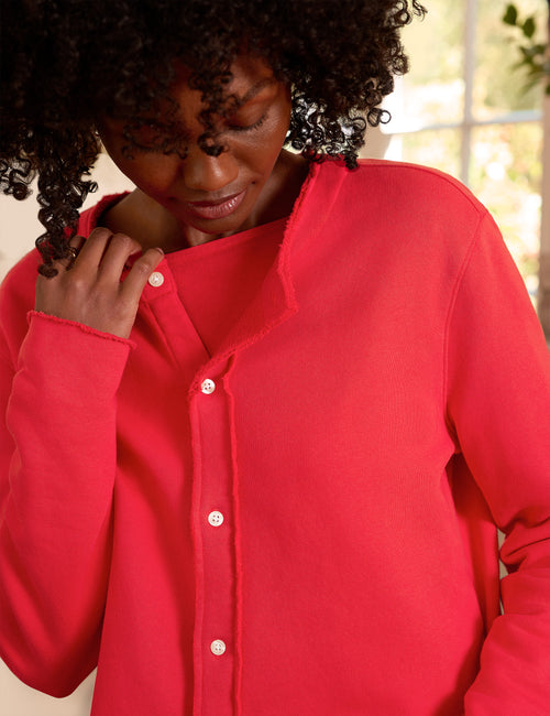 A close-up of a woman wearing a bright red long-sleeve button-up top, looking down and touching the collar area. The background is a softly blurred indoor setting with light-colored walls and a window., view 3