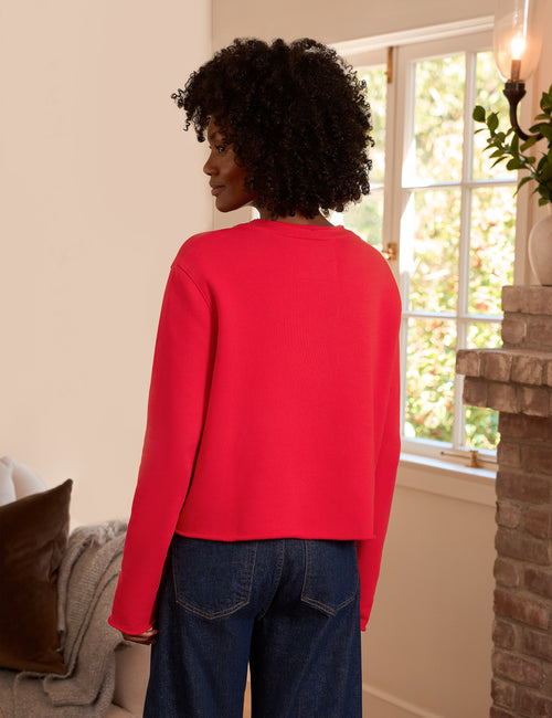 A woman with curly black hair is shown from behind wearing a bright red long-sleeve cropped sweater and dark blue jeans in a cozy indoor setting with a window and brick fireplace., alternate view