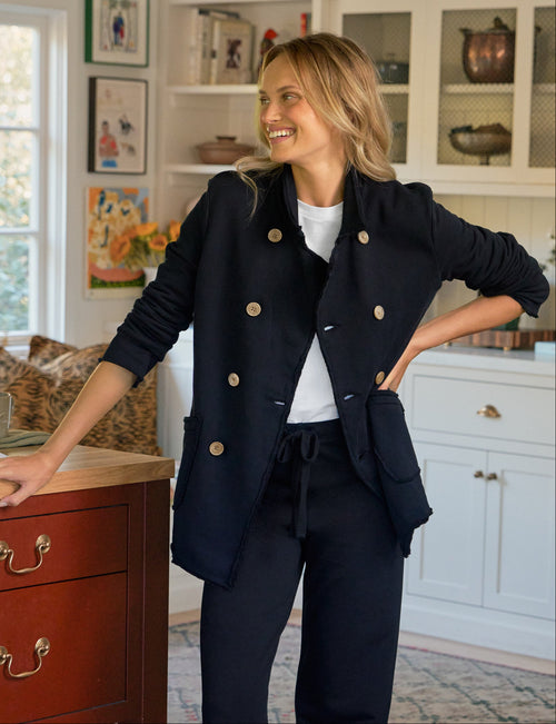 A woman stands in a bright home office wearing a tailored navy double-breasted blazer with matching trousers, a white top, and tan loafers. She is leaning against a wooden desk next to a window, with shelves and a patterned rug in the background., front view